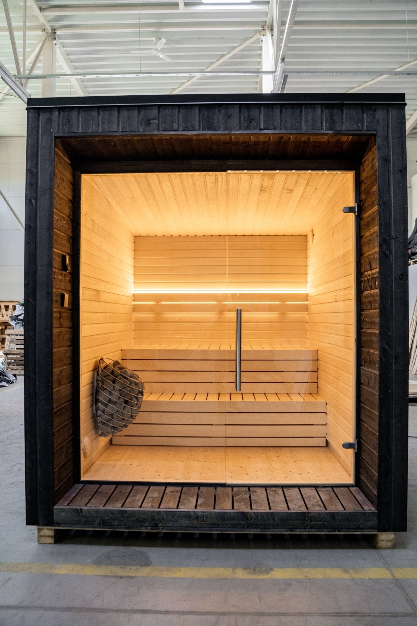 Sauna Exterior Lumi com porta de vidro e cesta de pedras.